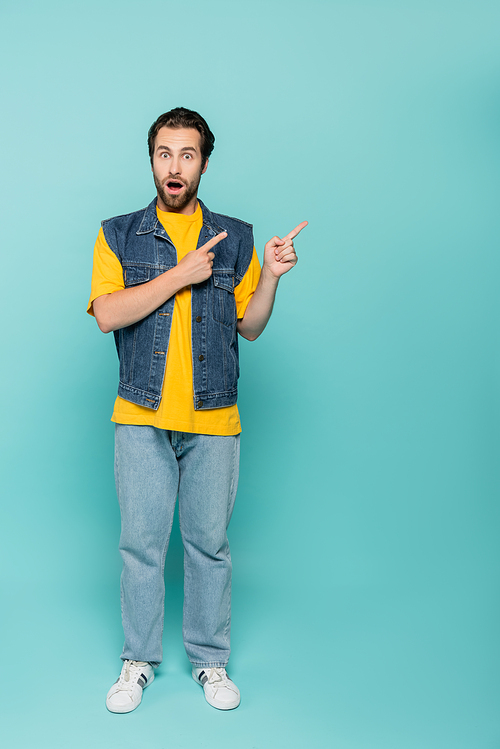 Amazed man in denim vest pointing with fingers on blue background
