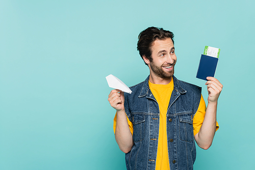 Smiling man holding paper plane and passport with air ticket isolated on blue