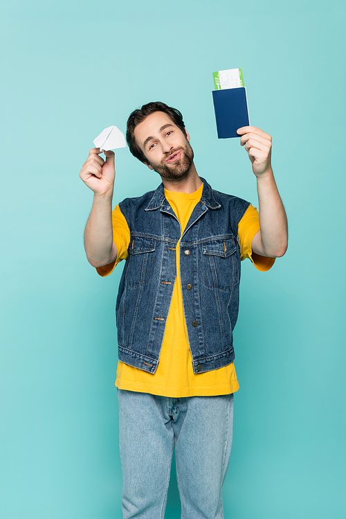Man pouting lips while holding paper plane and passport with air ticket isolated on blue
