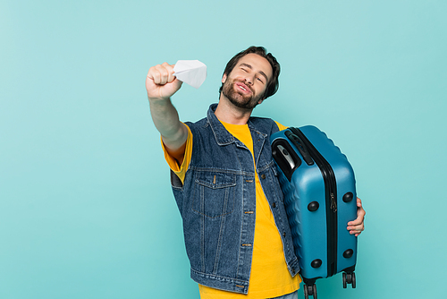 Positive man holding suitcase and playing with paper plane isolated on blue