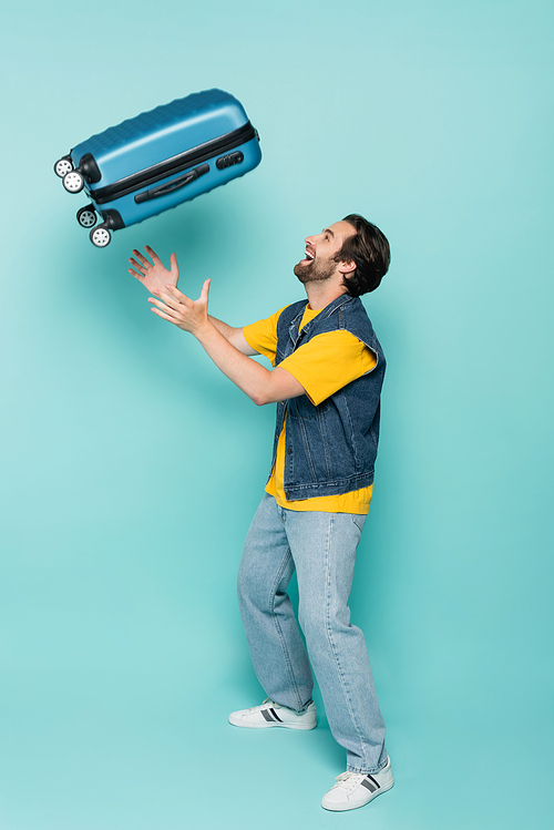 Side view of cheerful man throwing suitcase on blue background