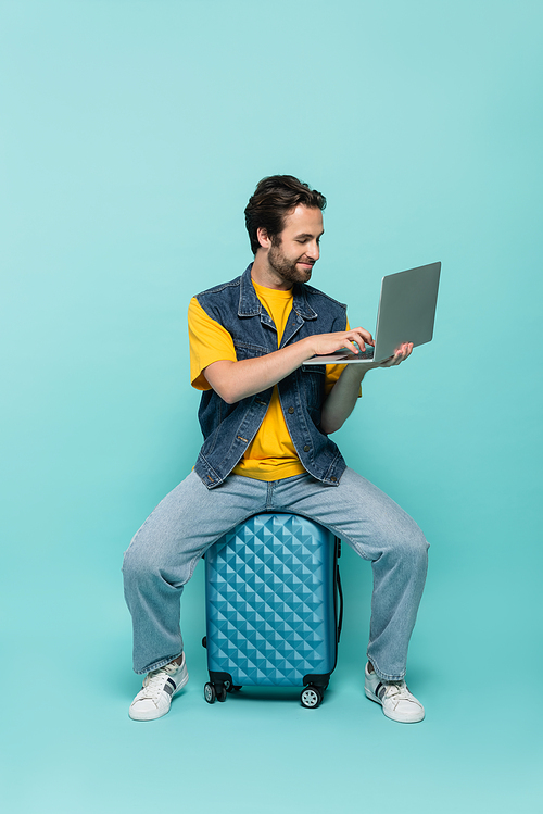 Smiling freelancer using laptop on suitcase on blue background