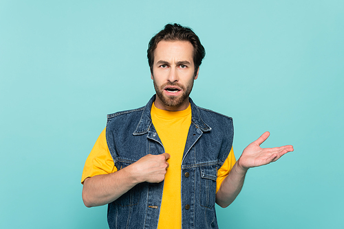 Confused man in denim vest pointing with finger isolated on blue