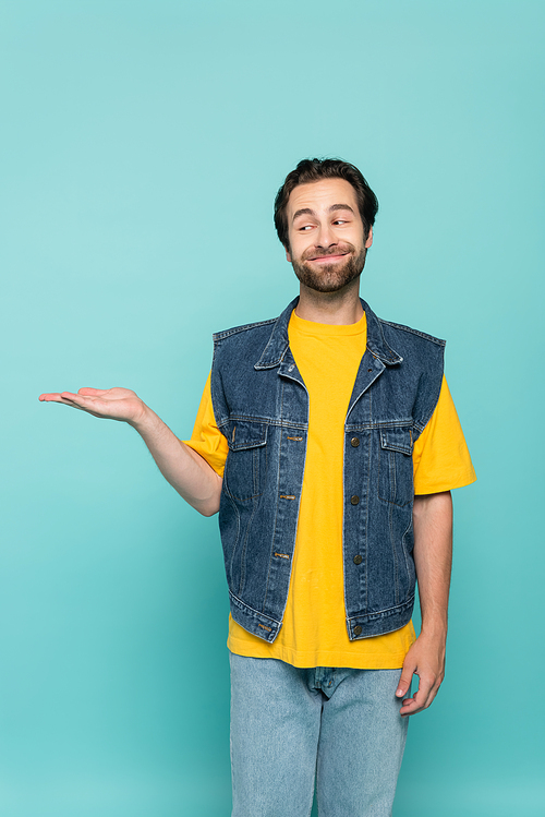 Smiling man in t-shirt and denim vest pointing with hand isolated on blue