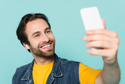 Smiling man in earphone taking selfie on smartphone isolated on blue