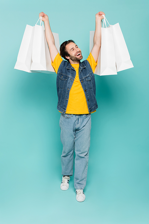 Full length of smiling man rising shopping bags on blue background