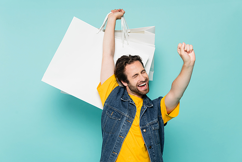 Happy man showing yes gesture and holding shopping bags isolated on blue