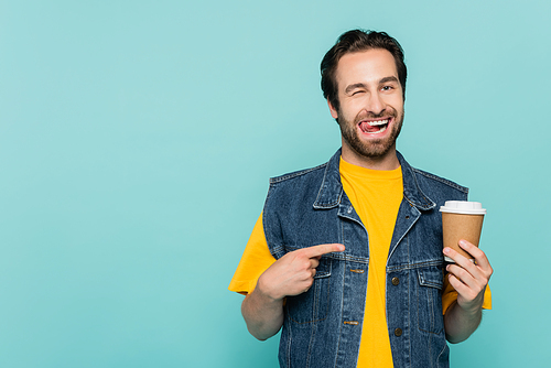 Young man sticking out tongue while pointing at coffee to go isolated on blue