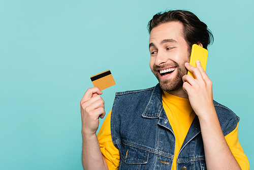 Smiling man talking on mobile phone and looking at credit card isolated on blue