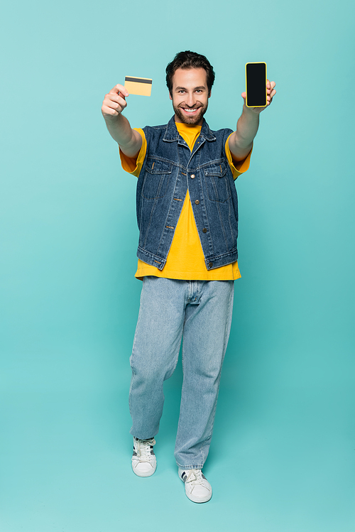 Positive man showing credit card and smartphone on blue background