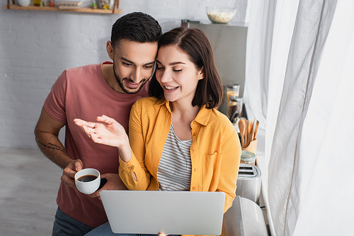 smiling young woman sitting with laptop and outstretched hand near boyfriend with coffee cup at home