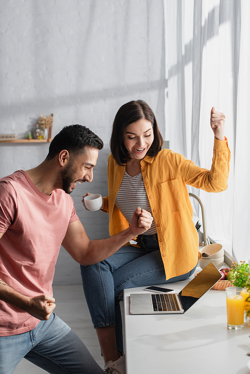 excited young couple looking at laptop with win gestures at home