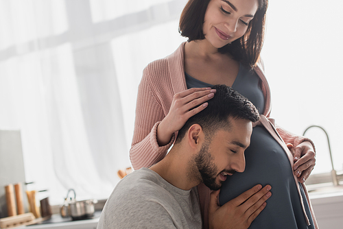 happy young man with closed eyes touching belly of pregnant woman in kitchen