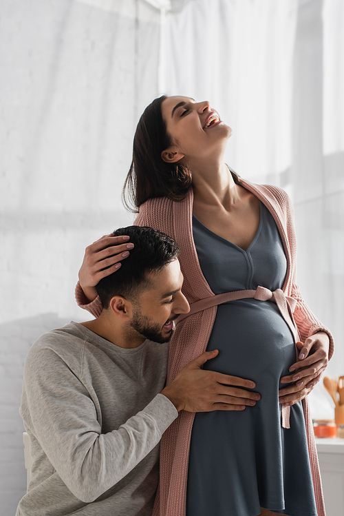 happy young man touching belly of pregnant woman in kitchen