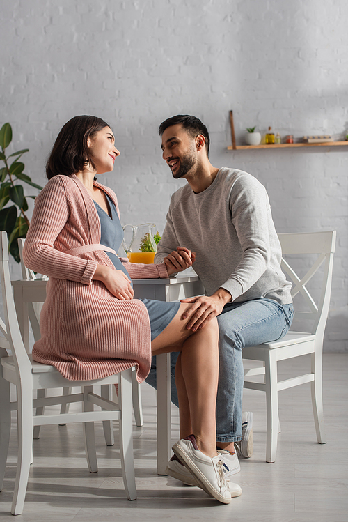 young man sitting near table and holding hand of pregnant woman in kitchen