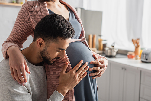 young man with closed eyes gently hugging belly of pregnant woman in kitchen