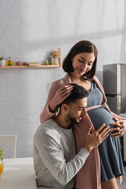 young man with closed eyes gently touching belly of pregnant woman in kitchen