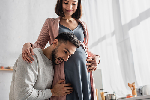 young man gently hugging pregnant woman and holding head near belly in kitchen