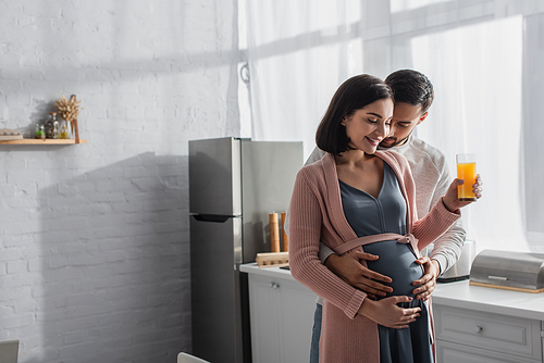 young man with closed eyes gently hugging pregnant woman with orange juice in kitchen