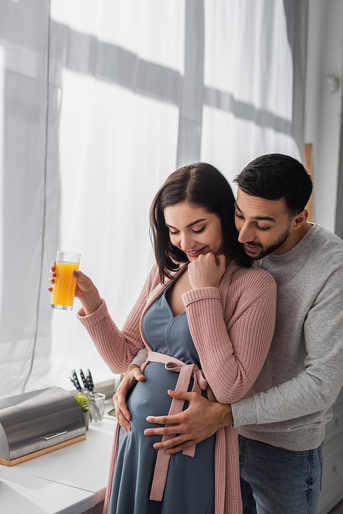 excited young man gently hugging pregnant woman with orange juice in kitchen