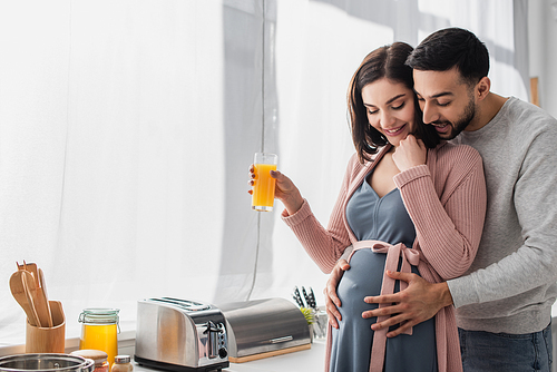 smiling young man gently hugging pregnant woman with orange juice in kitchen