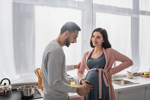 smiling young man gently touching pregnant woman in kitchen