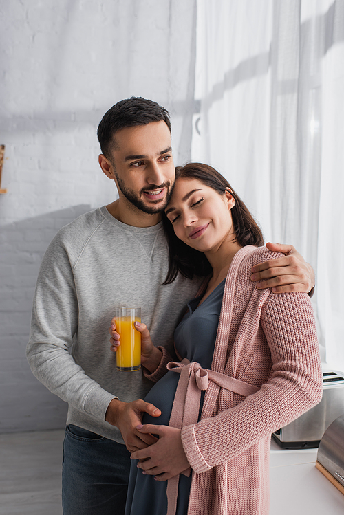 smiling young man hugging pregnant woman with orange juice in kitchen