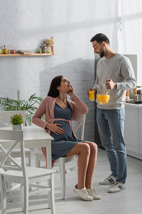 smiling young man standing with jar and glass of orange juice near pregnant woman in kitchen