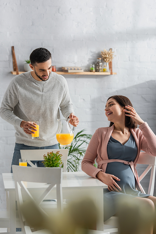 positive young man standing with jar and glass of orange juice near pregnant woman in kitchen