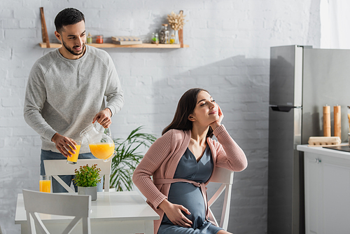 positive young man pouring orange juice from jar to glass near pregnant woman in kitchen