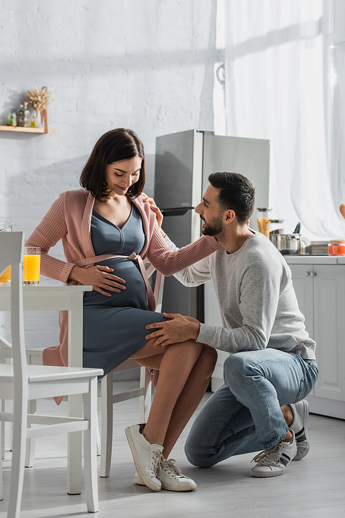 positive young man sitting on knee near pregnant woman in kitchen