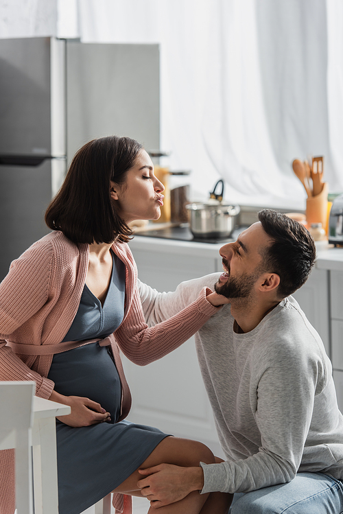 smiling young man sitting near pregnant woman with pouting lips in kitchen