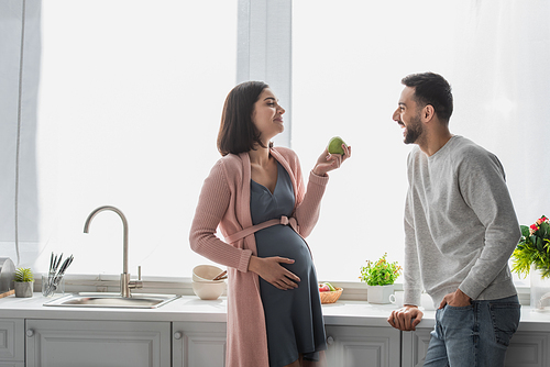 smiling young man standing near pregnant woman with apple in kitchen