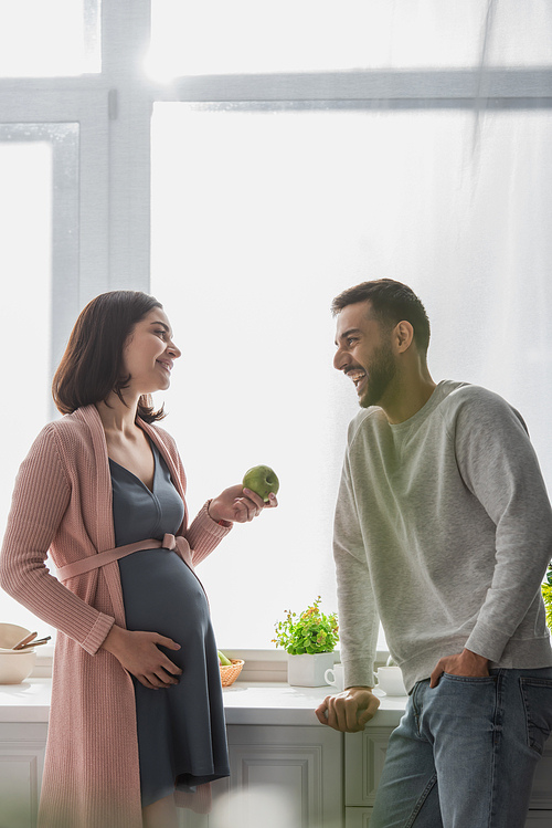 positive young man standing near pregnant woman with apple in kitchen