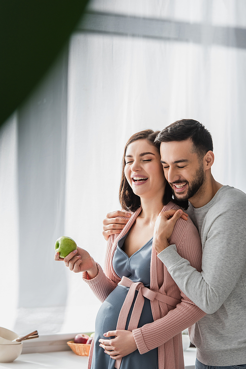 smiling young man with closed eyes hugging pregnant woman with apple in kitchen