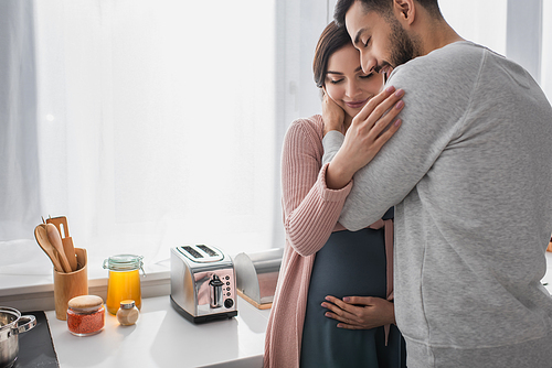 smiling young man with closed eyes hugging pregnant woman in kitchen