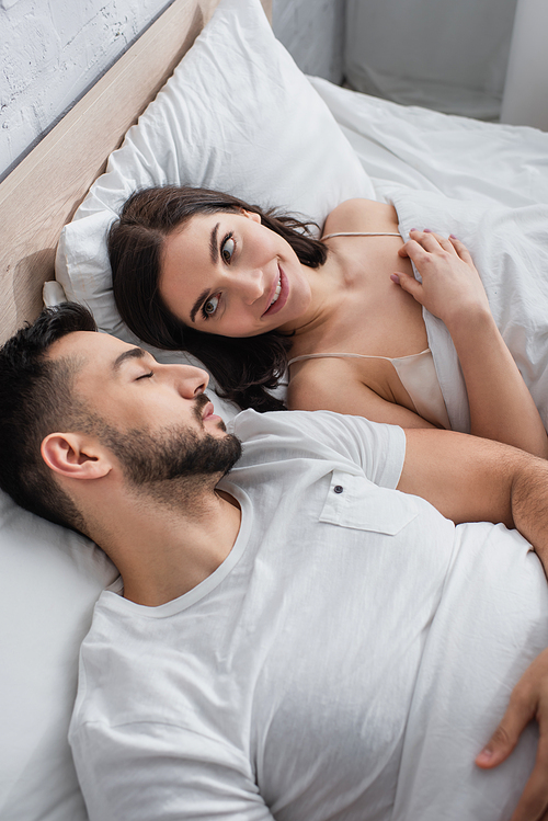 high angle view of smiling young woman looking at sleeping boyfriend in bed with white linen in bedroom