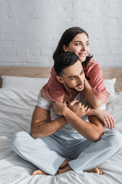 positive young couple sitting on bed with white linen and hugging in bedroom
