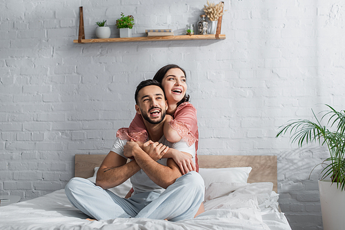 smiling young couple sitting on bed with white linen and hugging in bedroom