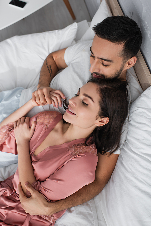 high angle view of smiling young couple lying on bed with white linen and holding hands in bedroom