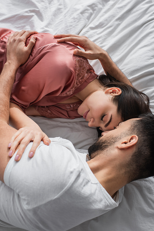 top view of young man lying on bed with white linen and gently hugging girlfriend with closed eyes in bedroom