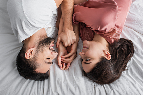 top view of young man lying on bed with white linen and gently holding hands of girlfriend in bedroom