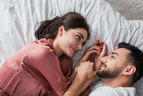 top view of smiling young couple lying on bed with white linen and looking at each other in bedroom