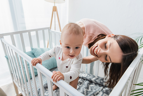 positive toddler child in romper standing in crib near happy mom