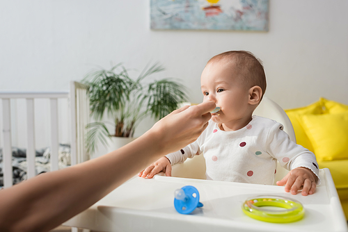 woman feeding little son near pacifier and rattle ring on baby chair