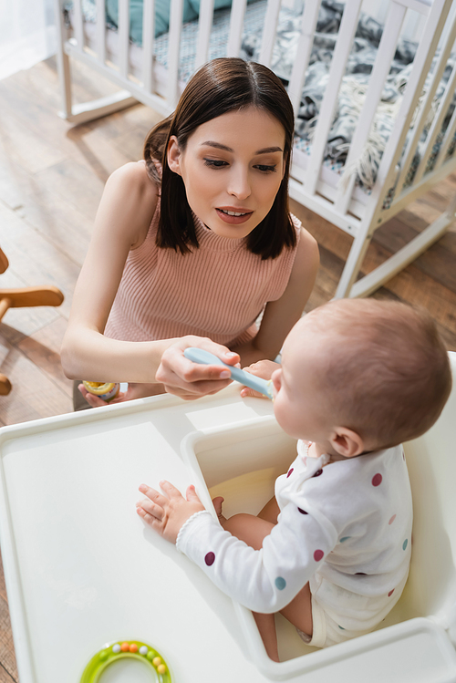 high angle view of brunette woman feeding blurred son in baby chair