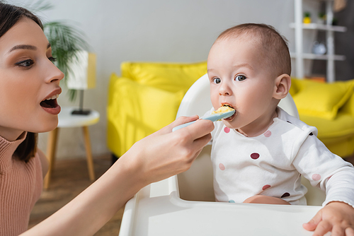brunette woman with open mouth feeding toddler kid in baby chair
