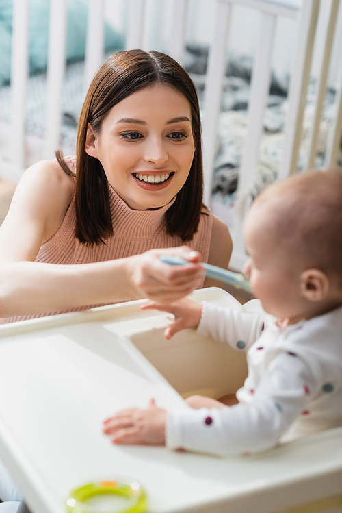 cheerful woman feeding blurred son from spoon at home