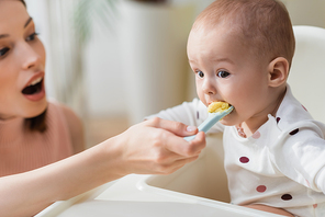 toddler kid in baby chair eating puree near blurred mother