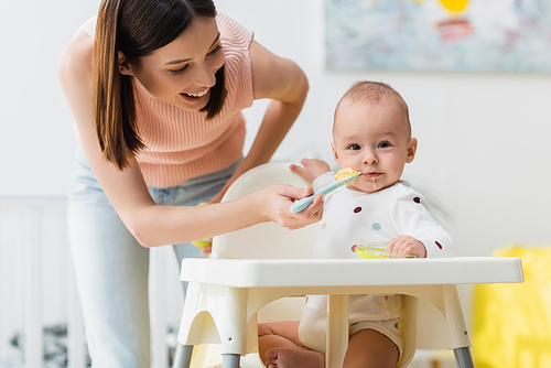 little boy in romper sitting in baby chair while happy mother feeding him from spoon